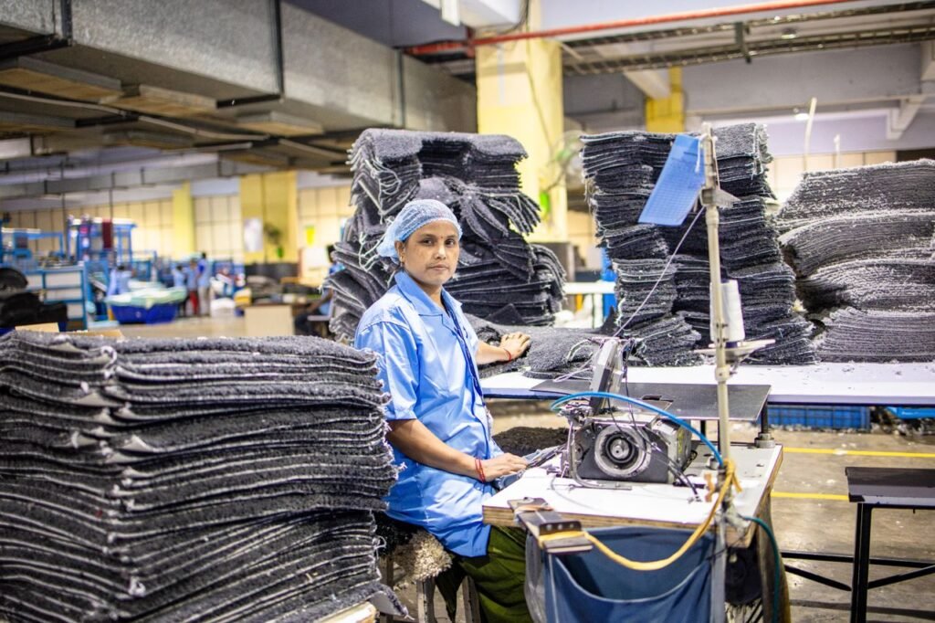 A factory worker operates a sewing machine surrounded by textile stacks in an industrial setting.
